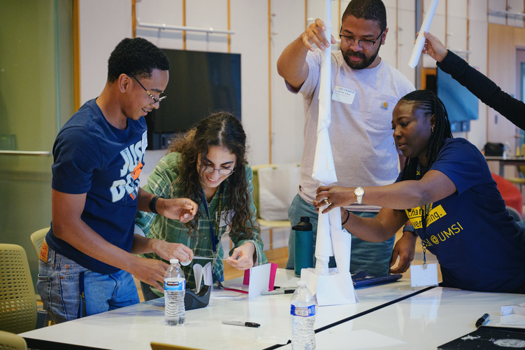 A team of five community college students work on creating a paper tower as a team building exercise.They reach their hands into the frame contributing pieces to the tower.