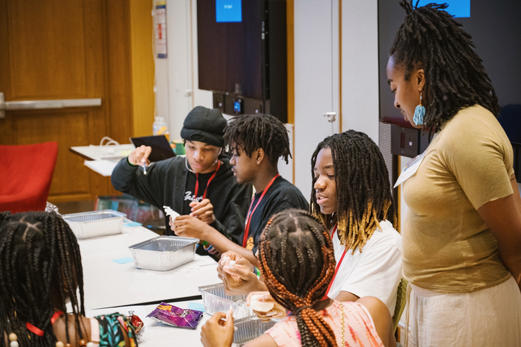 Five high school students sit at a table molding thermoplastics while a PhD student stands assisting them. 