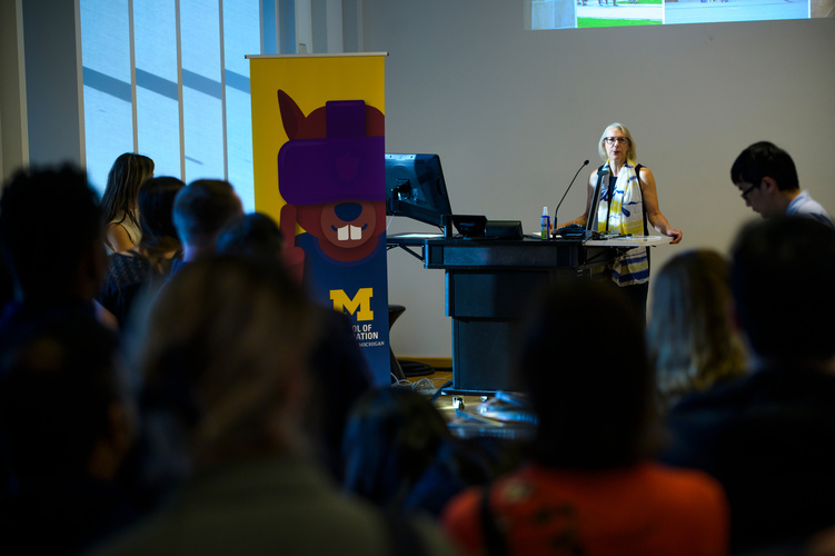 Interim dean Elizabeth Yakel stands at a podium facing a small crowd of students. To her right, there is a banner of UMSI's mascot, Mumsi (a squirrel wearing VR goggles).