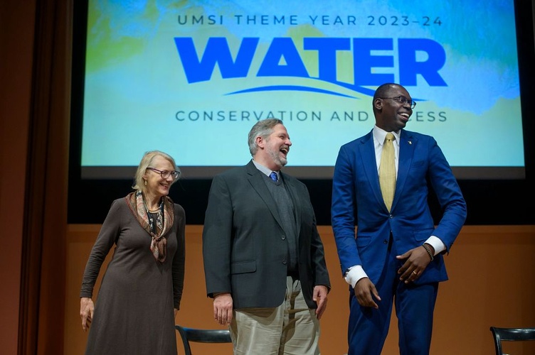 Elizabeth Yakel, Cliff Lampe and Lt. Gov. Garlin Gilchrist II smile on stage in front of a screen projecting "UMSI theme year 2023-24: Water conservation and access"