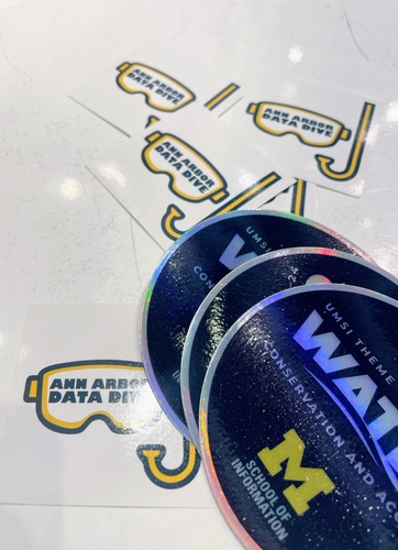 A photo of stickers that read "Ann Arbor Data Dive" alongside stickers with the UMSI theme year logo, which read "Water Conservation and Access"