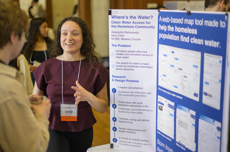 A student stands in front of a trifold poster, chatting animatedly with a guest at Exposition