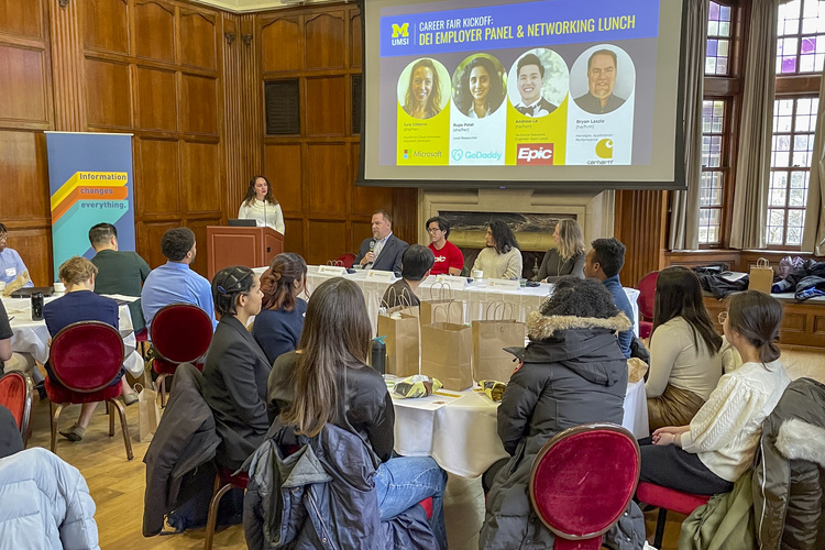 Employer representatives from Carhartt, Epic, Microsoft, and GoDaddy sit on a panel at the Michigan League to talk about their company’s DEI efforts and their own personal experiences. Students listen at round tables with bag lunches in front of them. 
