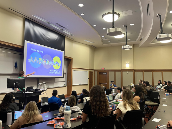 Representatives from Procter & Gamble present a slide about their brands to a classroom of students. In the foreground there are Procter & Gamble products the representatives brought as giveaways to students.
