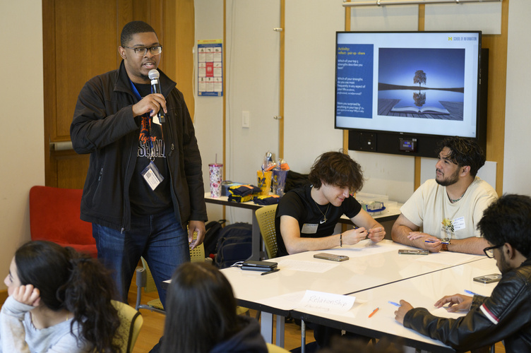 Marco Britton stands and speaks into a microphone, while others students sit at tables listening and taking notes. A screen behind him displays a presentation slide.