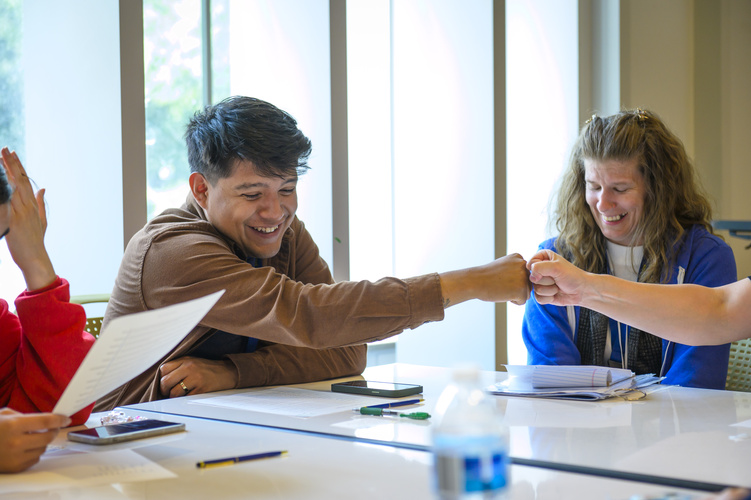 A student smiles while fist bumping another student who is just out of view. They are seated at a table with worksheets in front of them and sunny windows behind them.