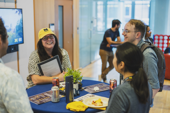 Dean Andrea Forte stands at a circular table with three other UMSI community members, laughing and wearing a yellow U-M cap. 