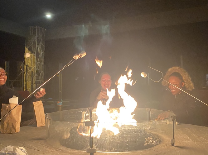 Three people laugh on the other side of a fire pit while holding up sticks to roast marshmallows