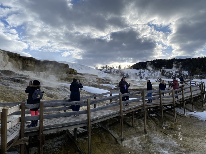 Seven traip members stand on a raised walkway beside Mammoth Hot Springs, each a few feet away from each other, a few looking through binoculars