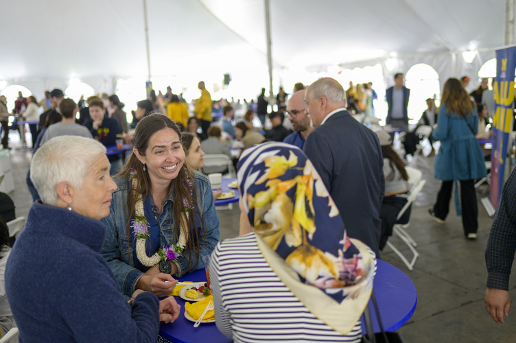 A person smiles while speaking to a small group at a blue table inside a bright white outdoor tent filled with people