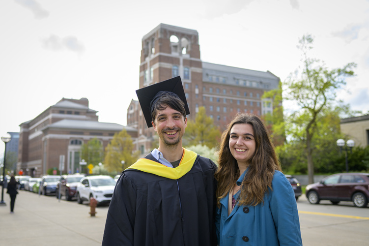 A graduate wearing a cap and gown poses beside a loved one wearing a blue jacket, with campus buildings and a road visible behind them
