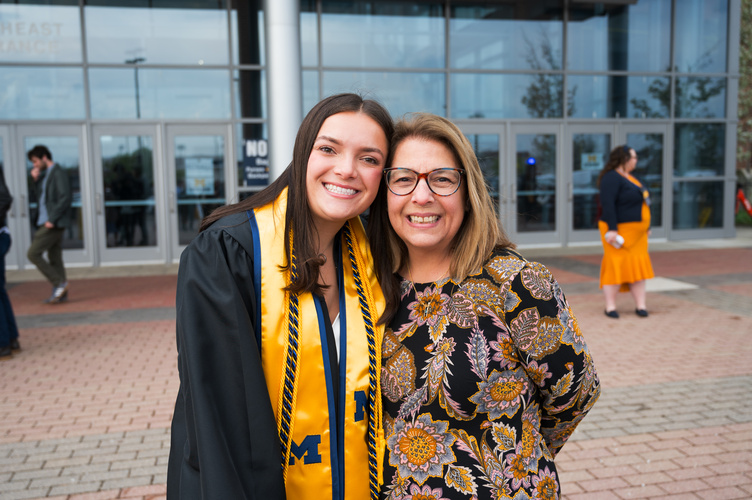 A graduate wearing a bright yellow stole poses with their head pressed against a family member's, whose smile resembles theirs.