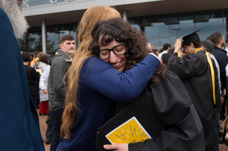 An graduate wearing glasses with a happy but emotional expression leans into a big hug from another person wearing blue 