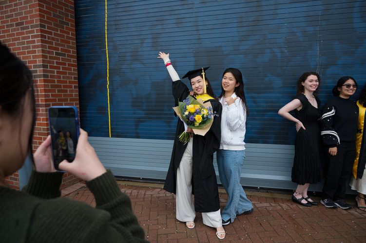 A graduate in a cap and gown, holding a yellow bouquet, gestures proudly with one arm up in the air, while posing beside another person in regular clothes. In the foreground, someone holds a phone out to take a photo of them.