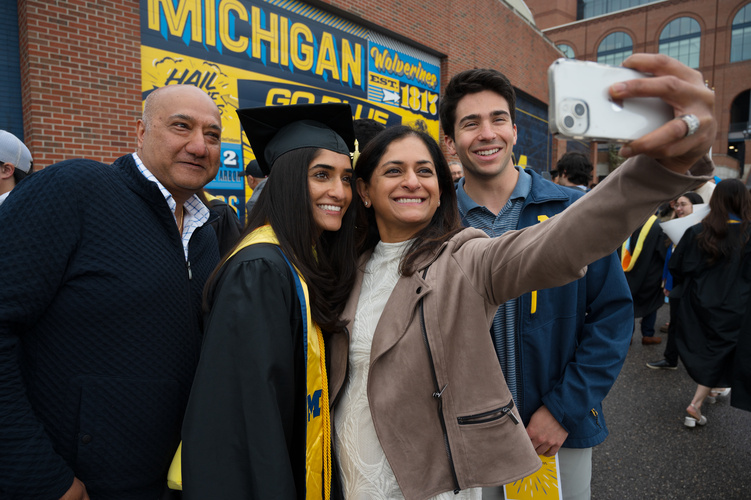 Four family members take a selfie, one of them in graduation garb, with a brick wall painted with Michigan branding visible behind them