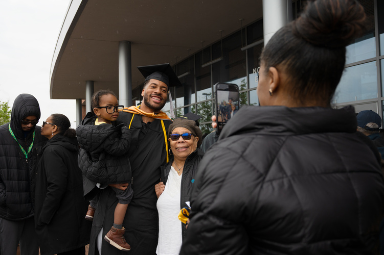 A graduate poses with family members while holding their small child, who is wearing oversized spectacles. Someone looks on from the foreground