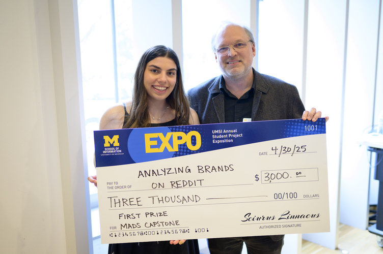 A graduate poses, smiling, beside Kevyn Collins-Thompson, holding a large award check with an Expo and UMSI logo
