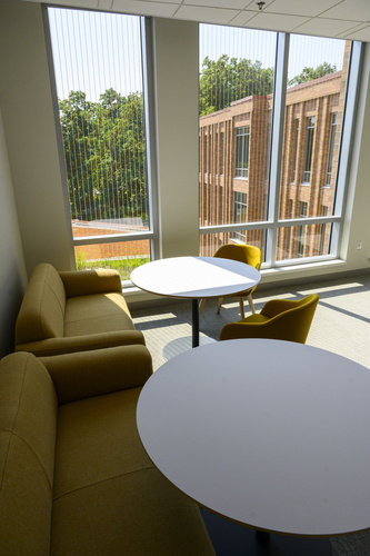 A close-up view of a nook in a building featuring yellow couches, round white tables, and yellow chairs beside tall windows allowing plenty of light in