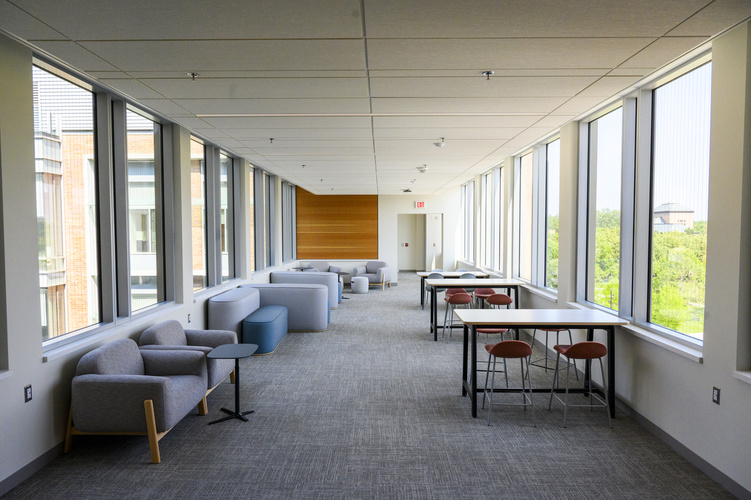 Interior of a modern lounge space featuring rows of tables with chairs and casual seating areas along large windows, with natural light and a view of the exterior greenery.
