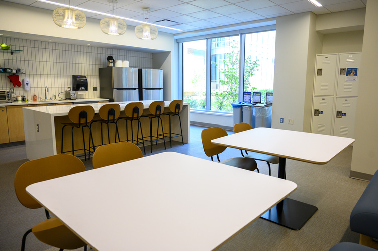 A modern kitchen with a long island lined with benches, white tables, two refrigerators, and a view of lockers in the right corner