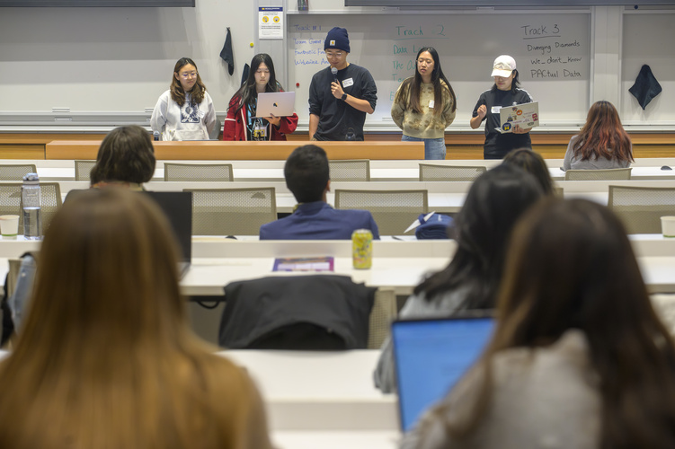 Five students stand in front of a classroom giving a presentation. Two hold laptops, while one holds a microphone