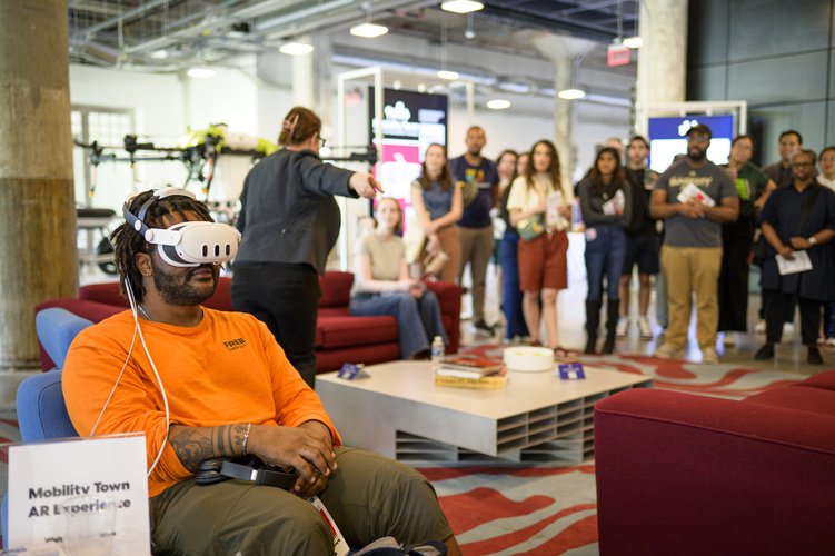 A student wears mixed reality goggles while a crowd looks on. 