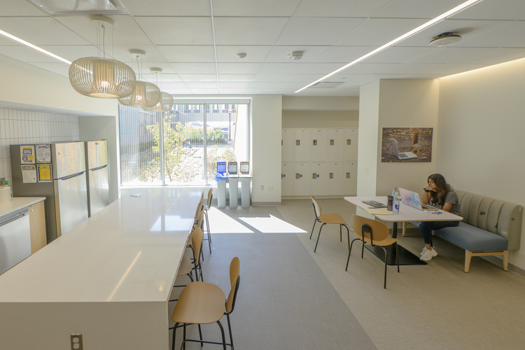 A wide view of the UMSI student lounge, with a large kitchen island, a row of white day-use lockers, and a single student seated at a table.