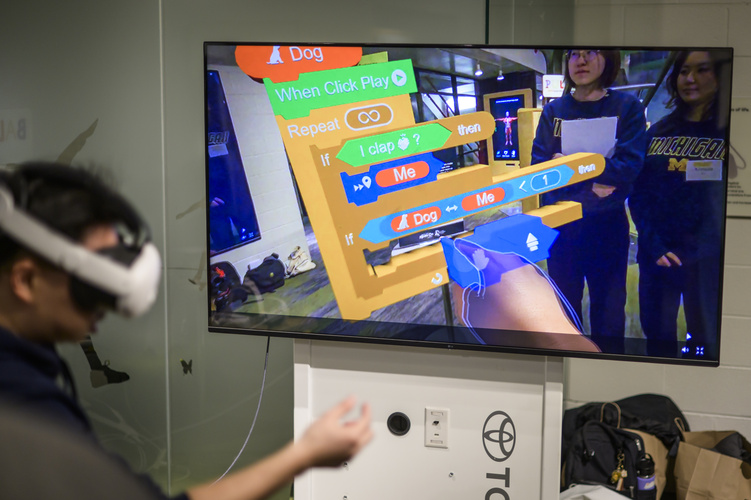 A person in a headset gestures. On a screen, a code sequence appears in colorful blocks.