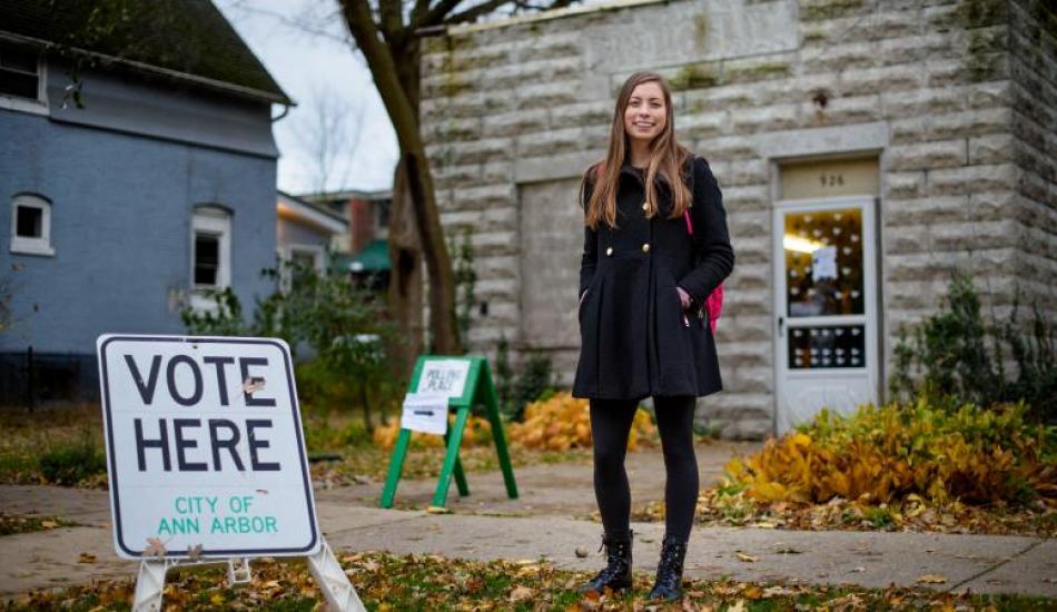 Shaelyn Albrecht standing in front of a polling location, there is a sign that says "vote here" to her right. 
