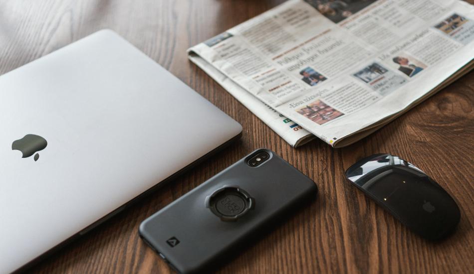 a laptop, a smartphone, a wireless mouse, and a newspaper arranged on a table