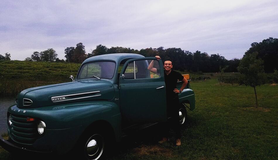 David Hernandez smiles and poses standing next to a vintage green pickup truck parked in a field under a light, cloudy sky.