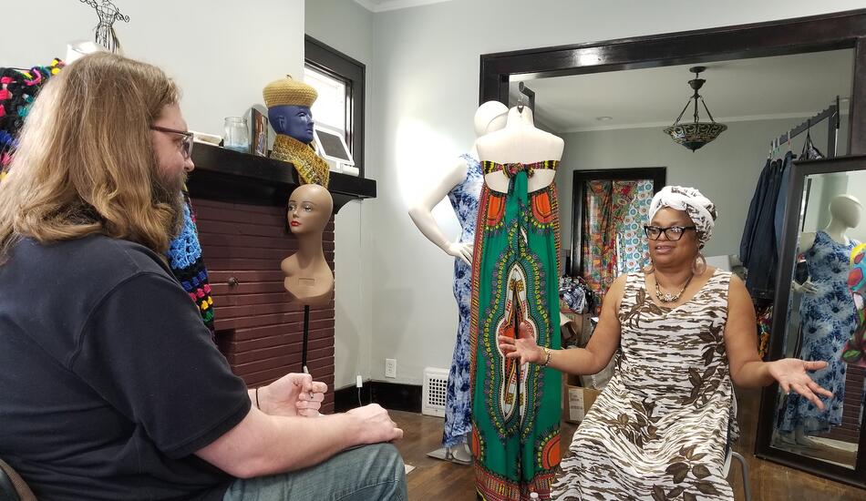 UMSI student Matt Garvin sits in a chair holding a pen, facing Duane Smith, who is speaking and gesturing. The room they are in has lots of mannequins wearing dresses and other clothing.