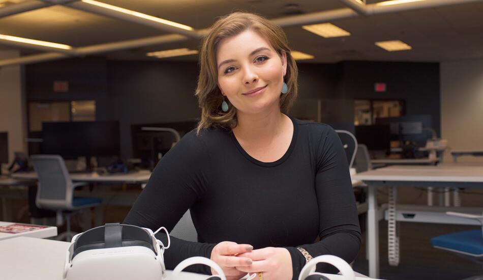 Chloe Preble smiles and sits at a table in the Visualization Study at the James and Ann Duderstadt Center. An Oculus headset and controllers lay on the table in front of her.