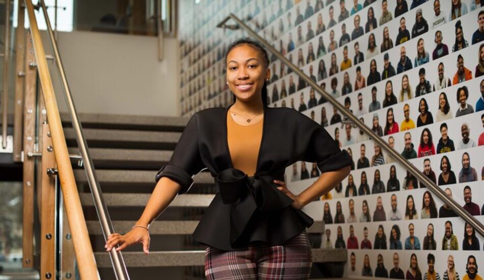 Morgan Miller stands and smiles on a staircase in the Trotter Multicultural Center, in front of a wall decorated with headshots of hundreds of U-M community members.