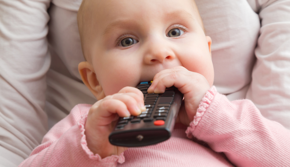Baby wearing a long-sleeve pink shirt and holding a remote to their mouth. 