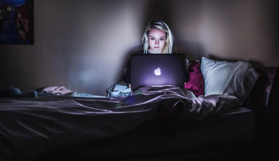 A person sits up in bed and uses a laptop in a dark room.