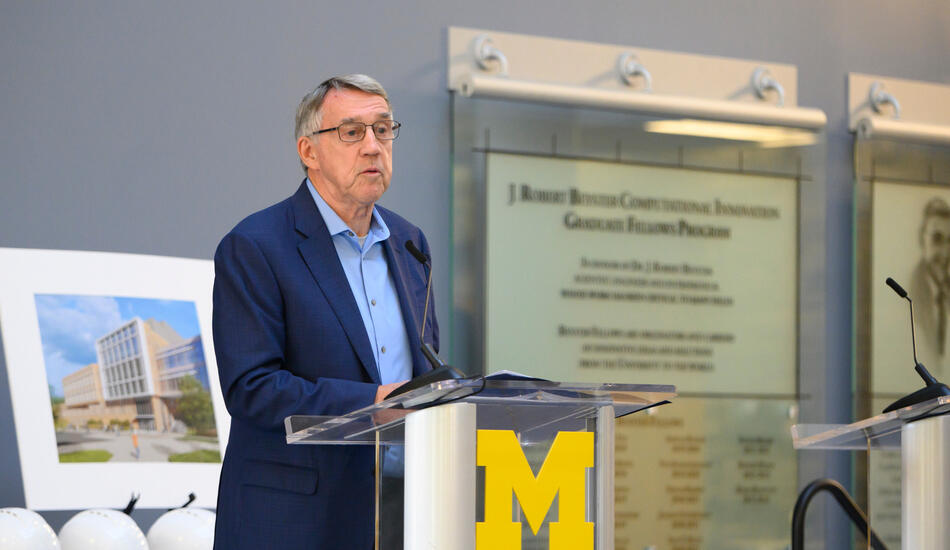 Donor Larry Leinweber speaks at a podium adorned with a Block M. Behind him are commemorative hardhats and a rendering of the Leinweber Computer Science and Information building. He is wearing glasses and a blue blazer.