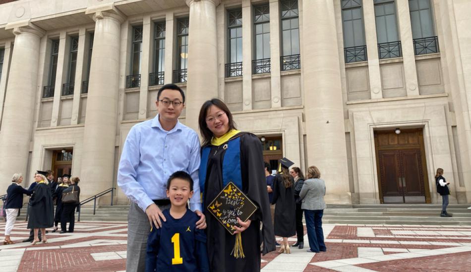 Yao Wei dressed in graduation gown stands outside of an auditorium on University of Michigan campus with her husband and son on her graduation day. She holds a decorated graduation cap.