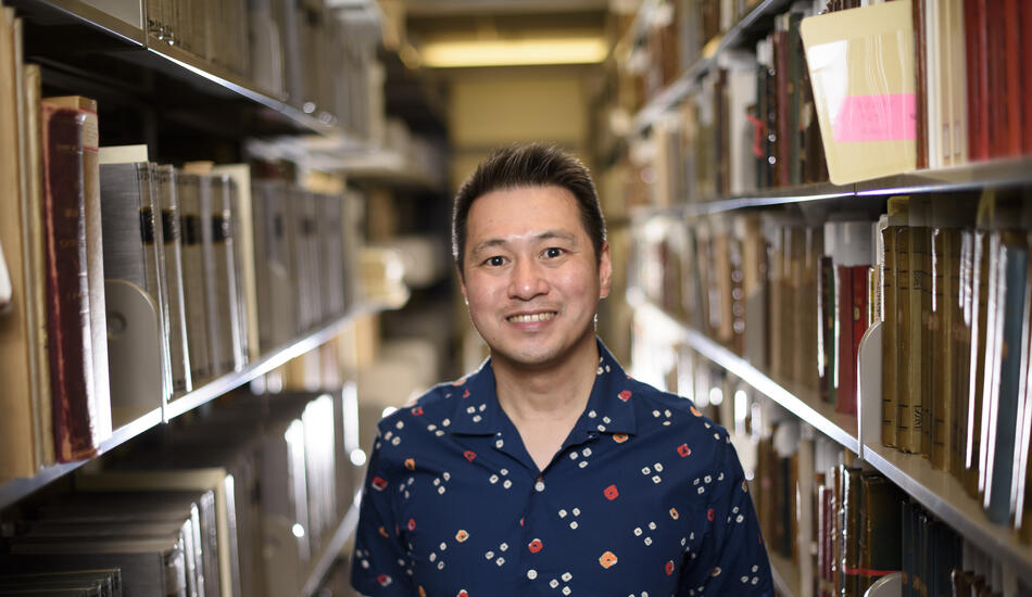 Ricky Punzalan, wearing a blue shirt, smiles as he stands among the stacks of archives in a University of Michigan library.