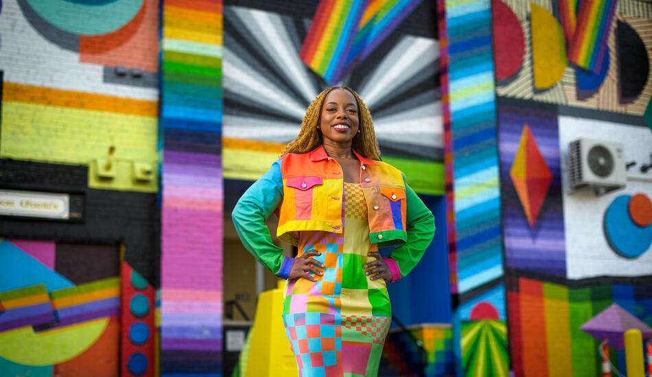 Megan poses in front of a rainbow and prismatic mural in an alleyway while wearing a vibrant dress and jacket of a patchwork of shapes and colors.