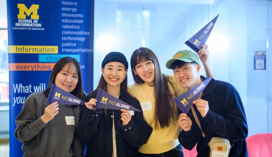 Four students holding pennants that say MADS alumni. They're standing in front of a UMSI banner.