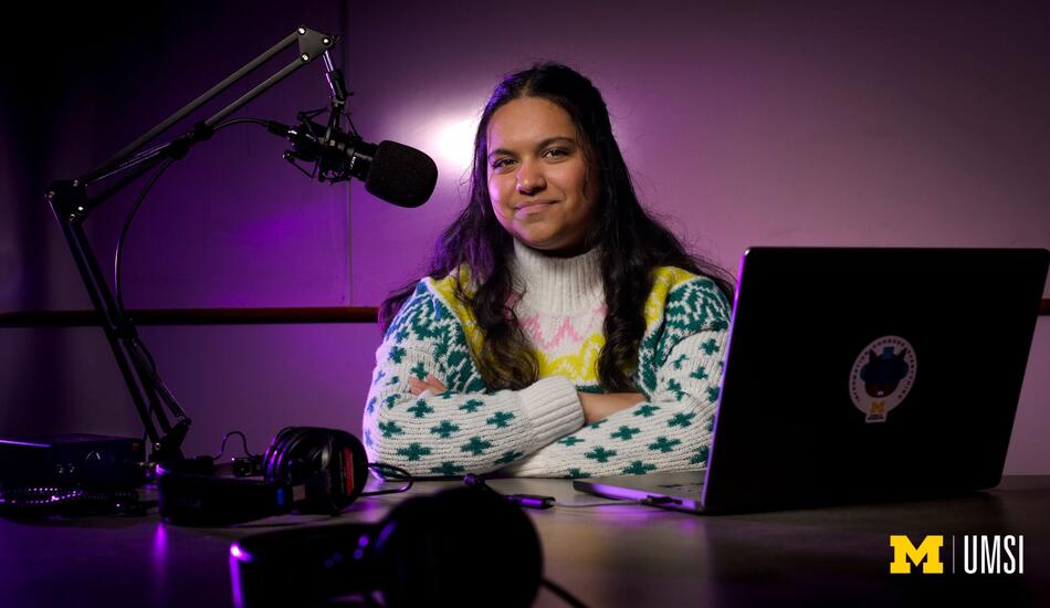 An image of Aayana Anand sitting in front of a podcasting microphone and computer with purple lighting behind her. 