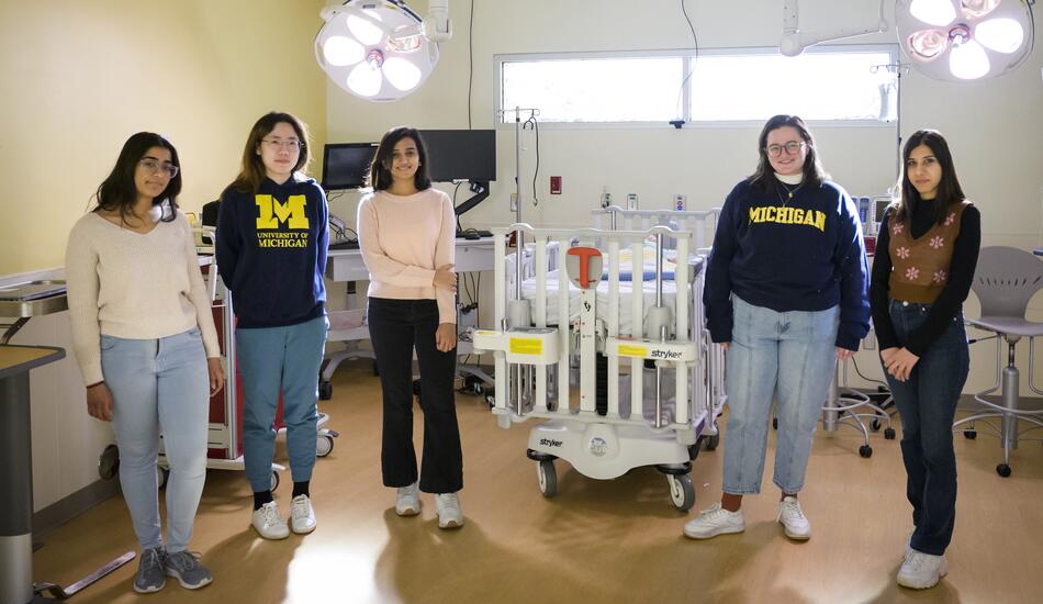A photo of five students standing in a light-filled operating room. Two of them wear "University of Michigan" sweatshirts.