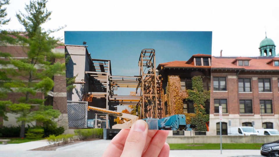 A hand holding a photo of a walkway being built between two buildings in front of those actual buildings.