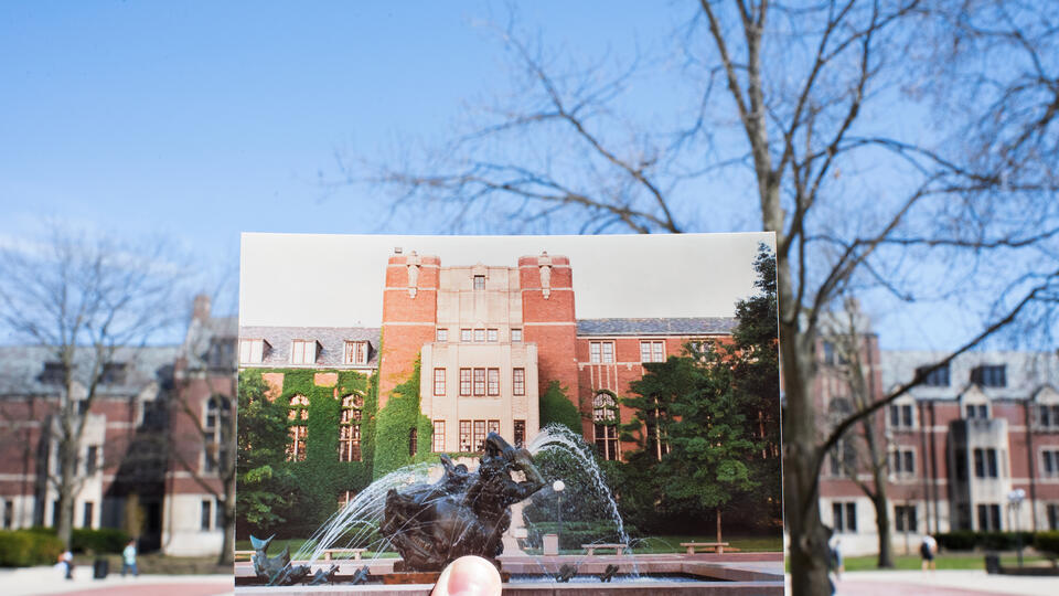 A hand holds a photo of a fountain and an ivy-covered building in front of the actual building. The only difference is that the ivy is gone now and the trees are green instead of bare.