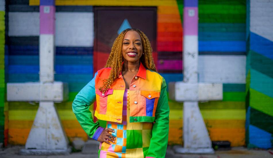 Megan poses in front of a rainbow and prismatic mural in an alleyway while wearing a vibrant dress and jacket of a patchwork of shapes and colors.