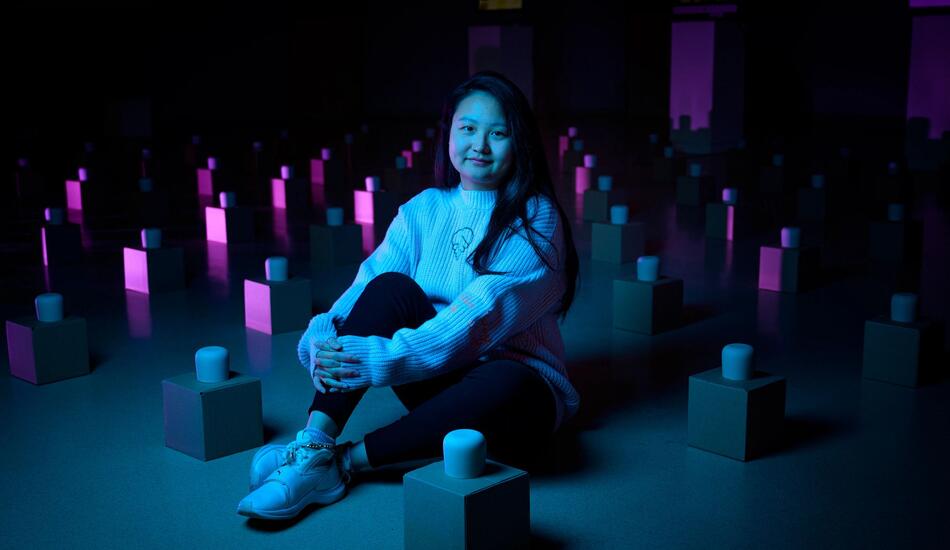 Lynette sits among an art exhibit of many tea cups on boxes laid out in a grid pattern across the floor.