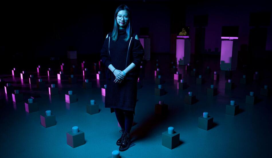 Jessie stands among an art exhibit of many tea cups on boxes laid out in a grid pattern across the floor.