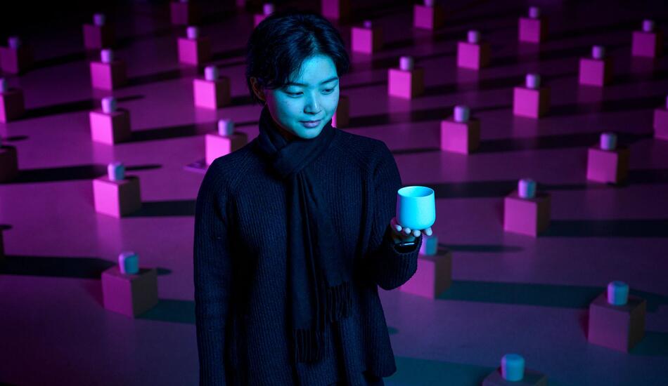 Lynette sits among an art exhibit of many tea cups on boxes laid out in a grid pattern across the floor.