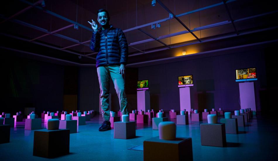 Dev holds up a peace sign and stands among an art exhibit of many tea cups on boxes laid out in a grid pattern across the floor.